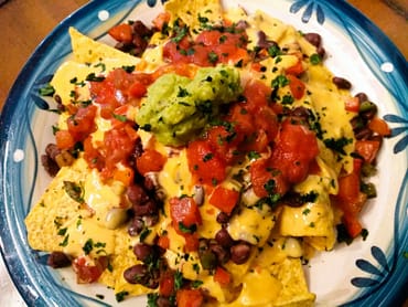 overhead yellow corn nacho chips covered in yellow nacho cheese, black beans, red salsa, peppers, onions, and topped with green guacamole