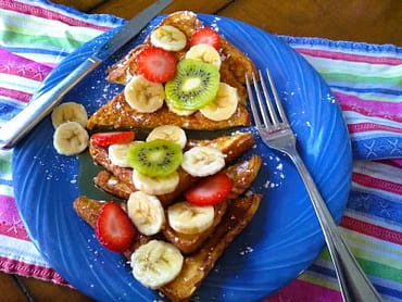 brioche french toast topped with bananas, kiwi, and strawberries, on a blue plate and colorful cloth.