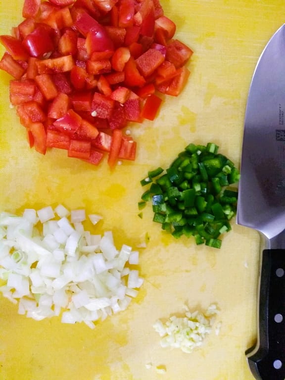 Diced red bell pepper, jalapeno, onion, and garlic on yellow cutting board with chef's knife